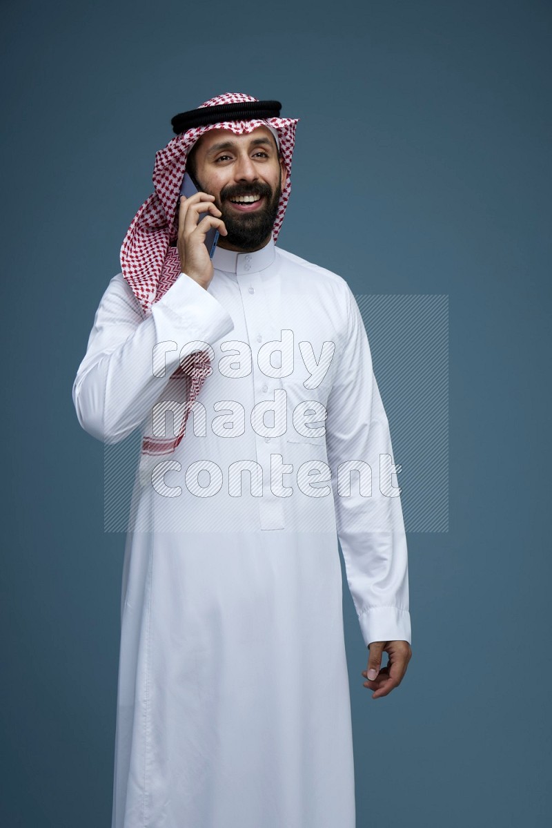 A man having a Call in a blue background wearing Saudi Thob with Shomag