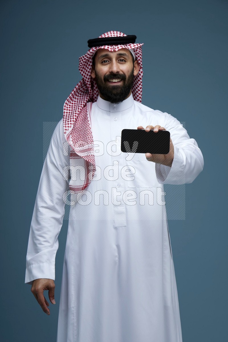 A man showing his phone Screen in a blue background wearing Saudi Thob with Shomag