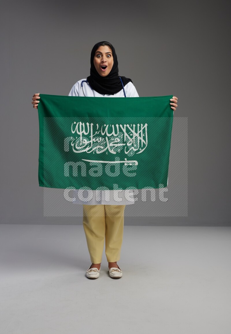 Saudi woman wearing lab coat with stethoscope standing holding Saudi flag on Gray background