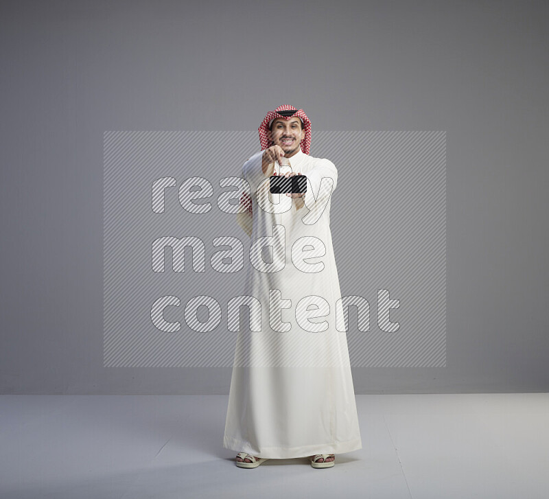 رجل سعودي يرتدي ثوب ابيض وشماغ احمر يشير الى هاتفه باتجاه الكاميرا