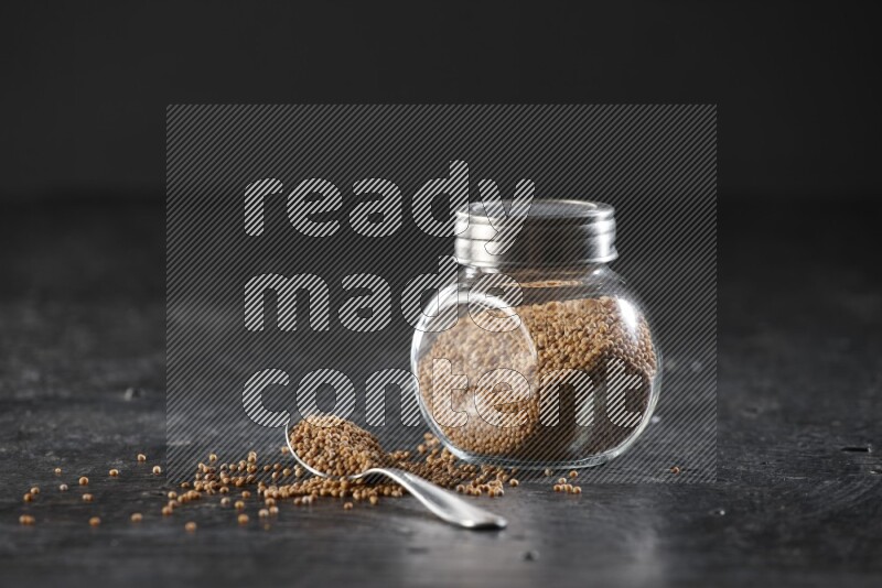 A glass spice jar and a metal spoon full of mustard seeds on a textured black flooring