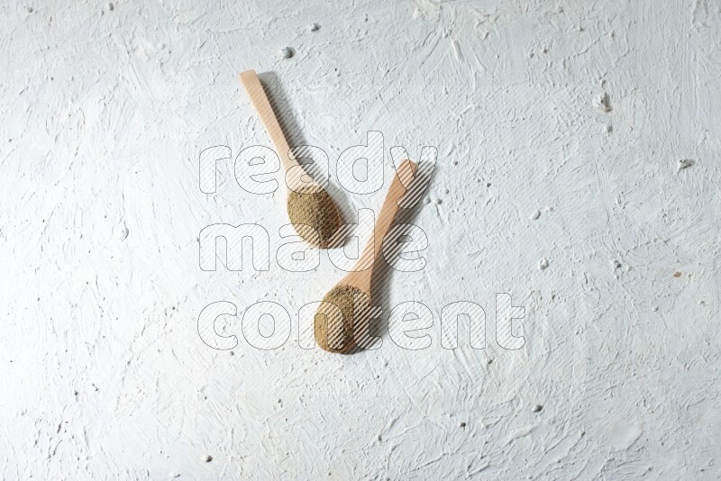 2 Wooden spoons full of cumin powder on a textured white flooring