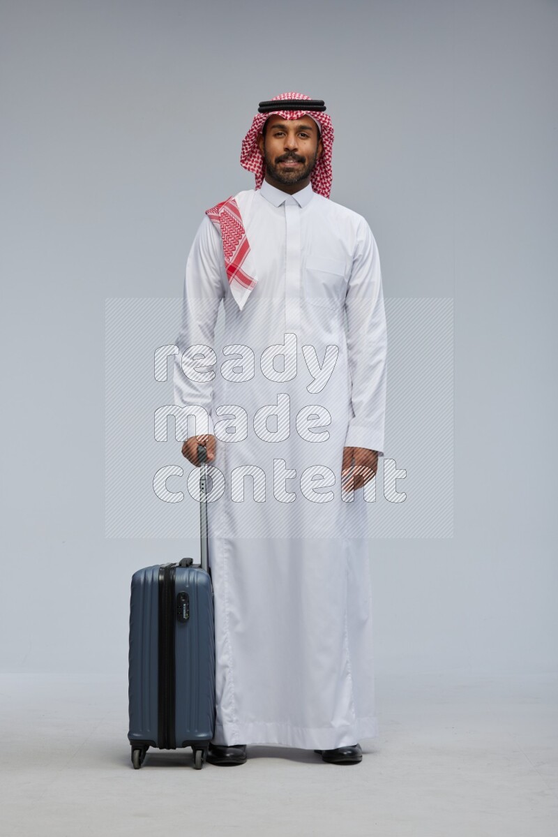 Saudi man wearing Thob and shomag standing holding Travel bag on Gray background