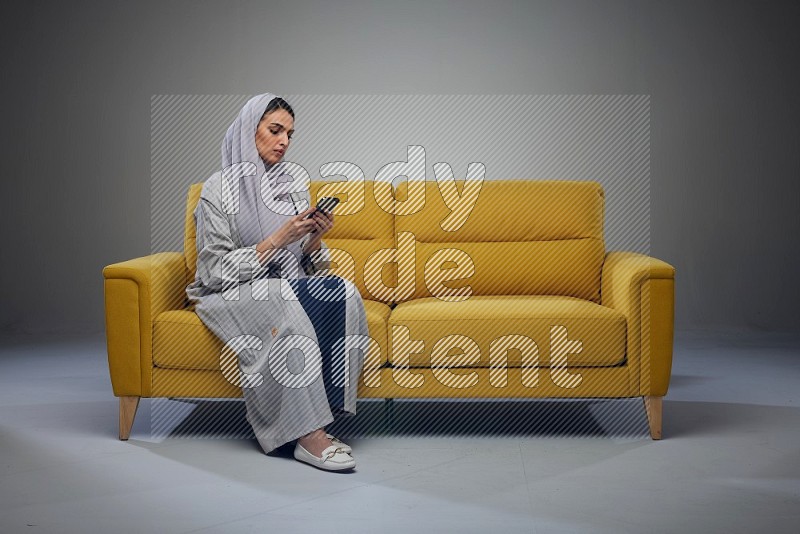 A Saudi woman wearing a light gray Abaya and white head scarf sitting on a yellow sofa and using her phone eye level on a grey background