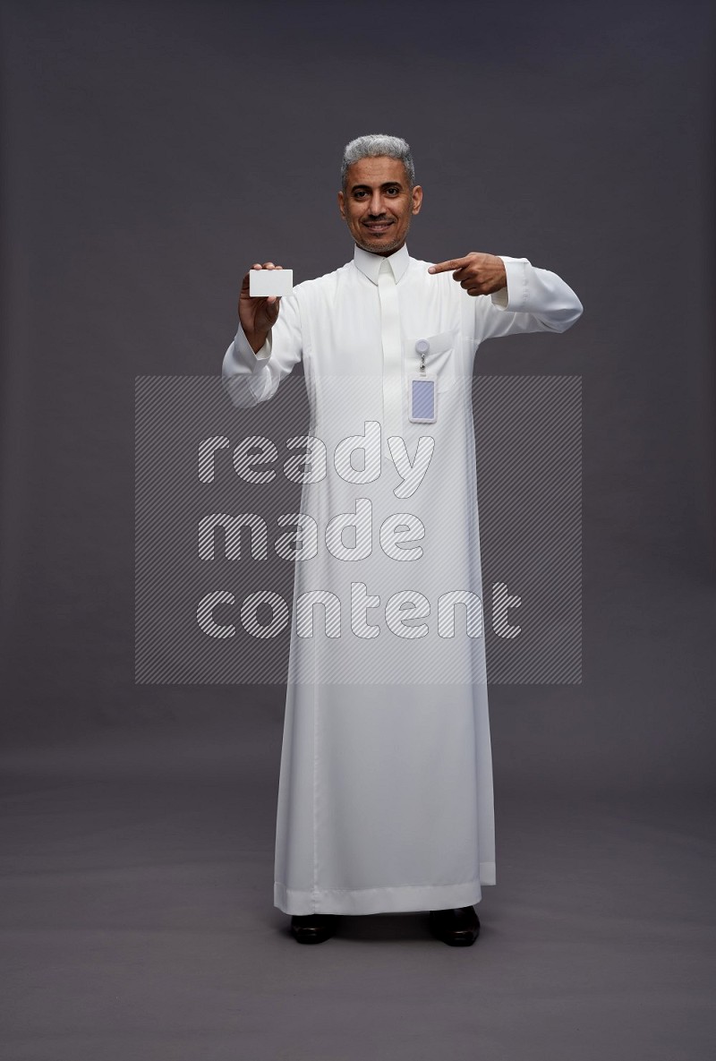 Saudi man wearing thob with pocket employee badge standing holding ATM card on gray background