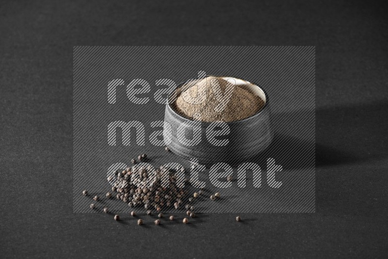 A black pottery bowl full of black pepper powder and black pepper beads on the floor on black flooring