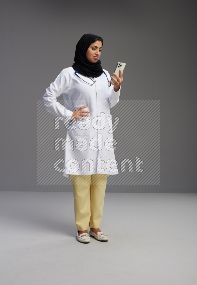 Saudi woman wearing lab coat with stethoscope standing texting on phone on Gray background