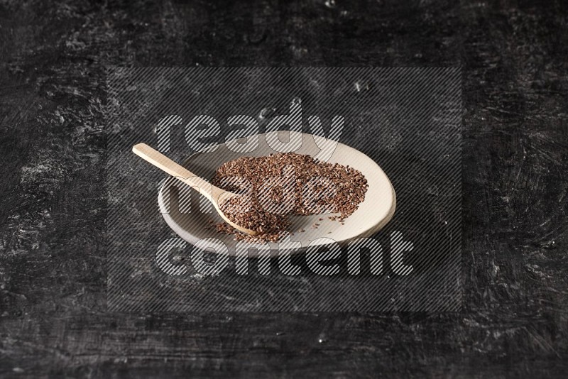 طبق فخار متعدد الألوان ممتلئ بحبوب بذر الكتان وملعقة خشبية ممتلئة بالبذور على أرضية سوداء