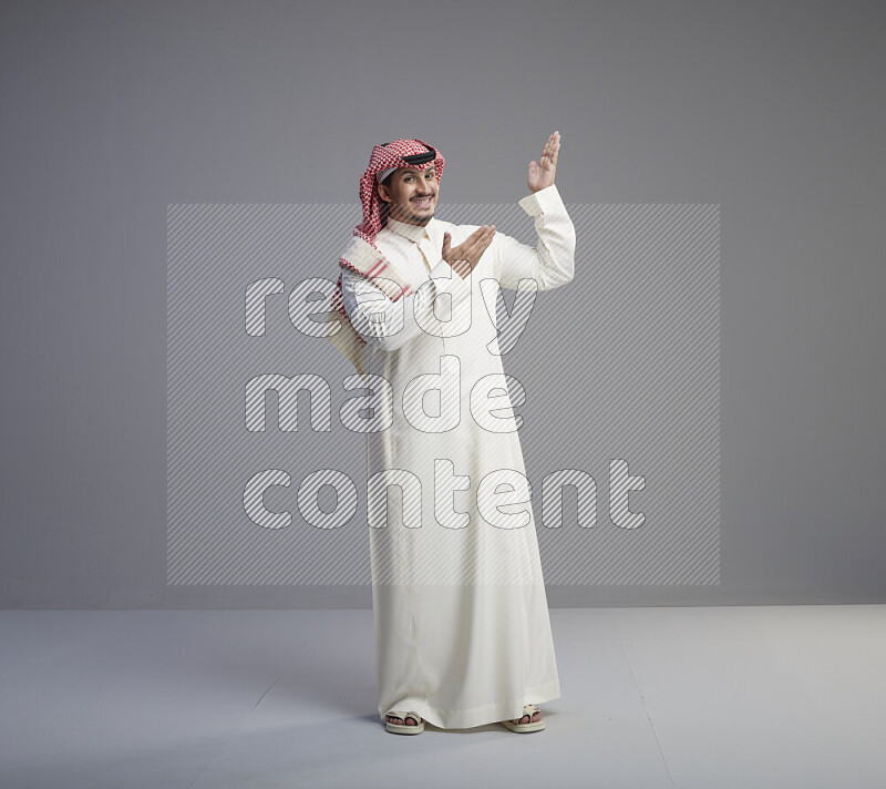 A Saudi man standing wearing thob and red shomag interacting with the camera on gray background