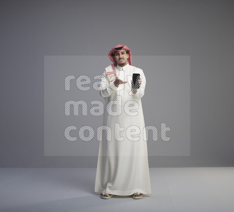 A Saudi man standing wearing thob and red shomag showing phone to camera on gray background
