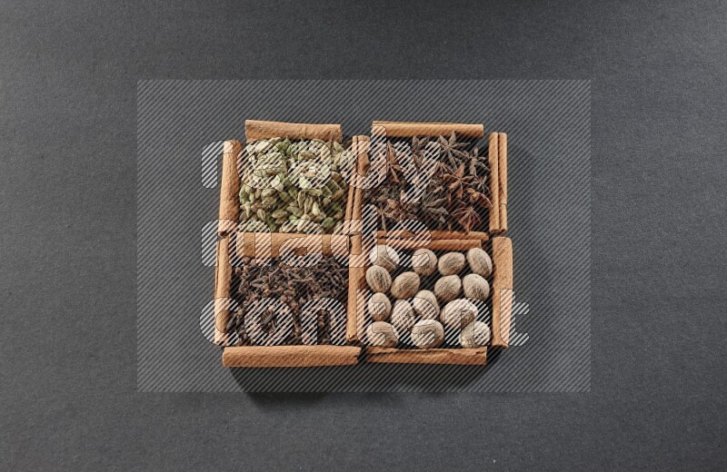 4 مربعات من عيدان القرفة ممتلئة بحب الهيل واليانسون النجمي وجوزة الطيب والقرنفل على أرضية سوداء