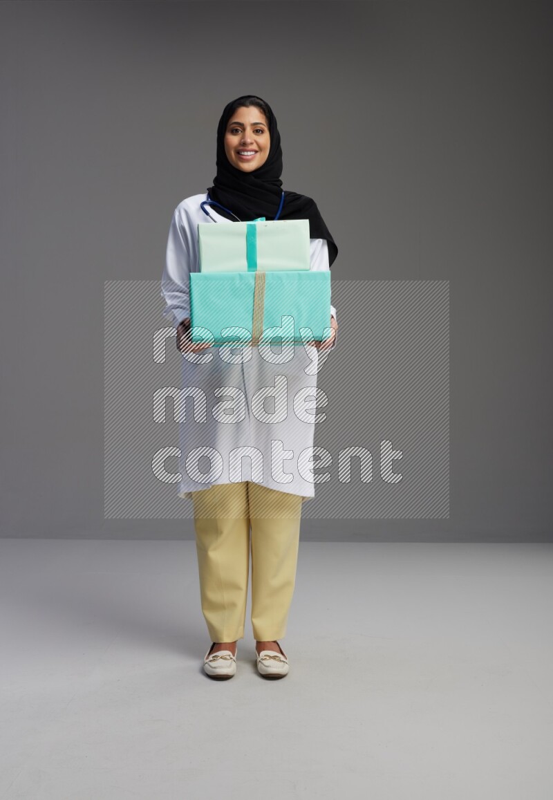 Saudi woman wearing lab coat with stethoscope standing holding gift box on Gray background