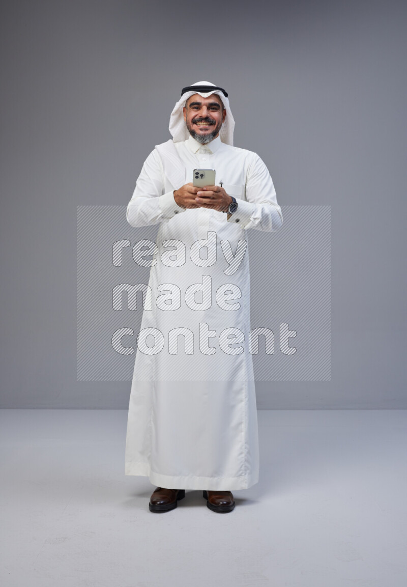 Saudi man Wearing Thob and white Shomag standing texting on phone on Gray background