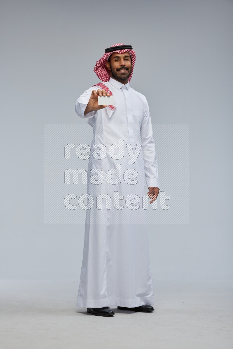 Saudi man wearing thob and shomag standing holding ATM card on gray background