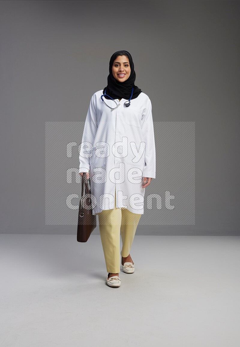 Saudi woman wearing lab coat with stethoscope standing holding bag on Gray background
