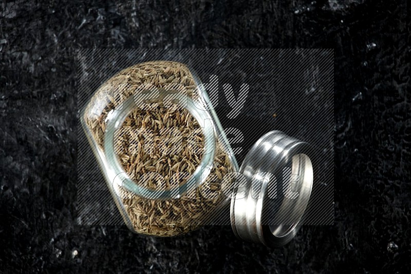 A glass spice jar full of cumin seeds on a textured black flooring