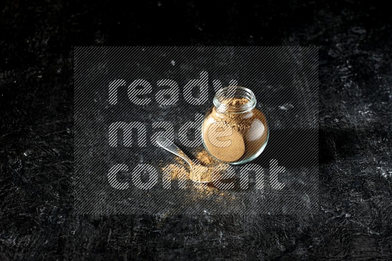 A glass spice jar and metal spoon full of allspice powder on a textured black flooring