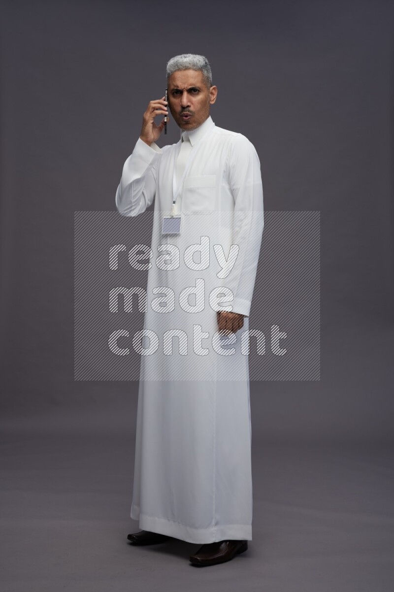 Saudi man wearing thob with neck strap employee badge standing talking on phone on gray background