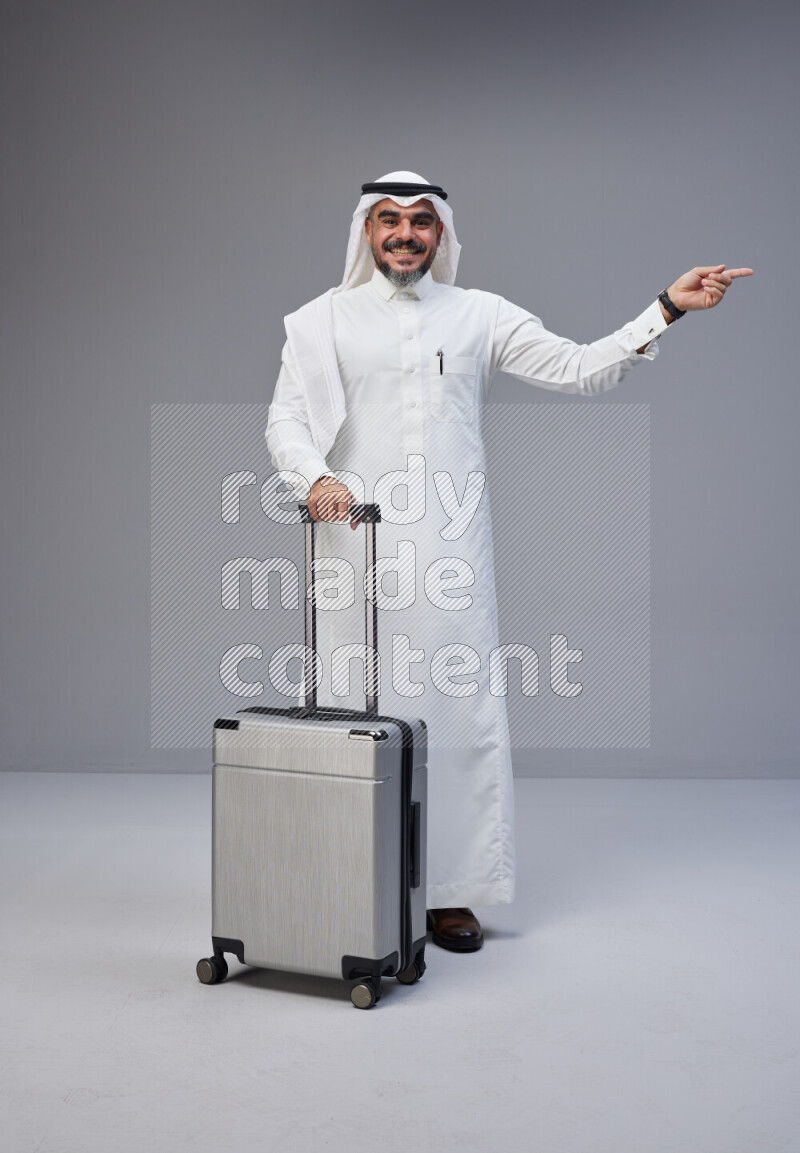 Saudi man wearing Thob and white Shomag standing holding Travel bag on Gray background