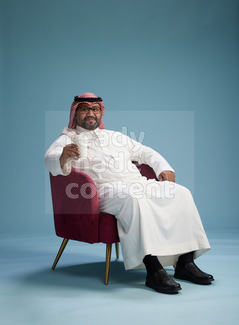 Saudi Man with shimag sitting on chair drinking coffee on blue background