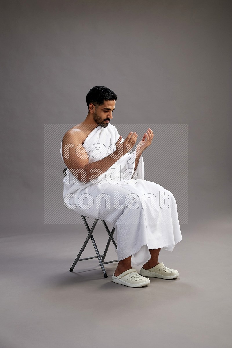 A man wearing Ehram sitting on chair performing dua'a on gray background