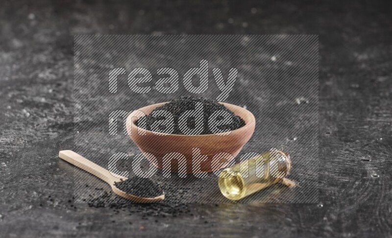A wooden bowl and spoon full of black seeds and a bottle of black seeds oil on a textured black flooring