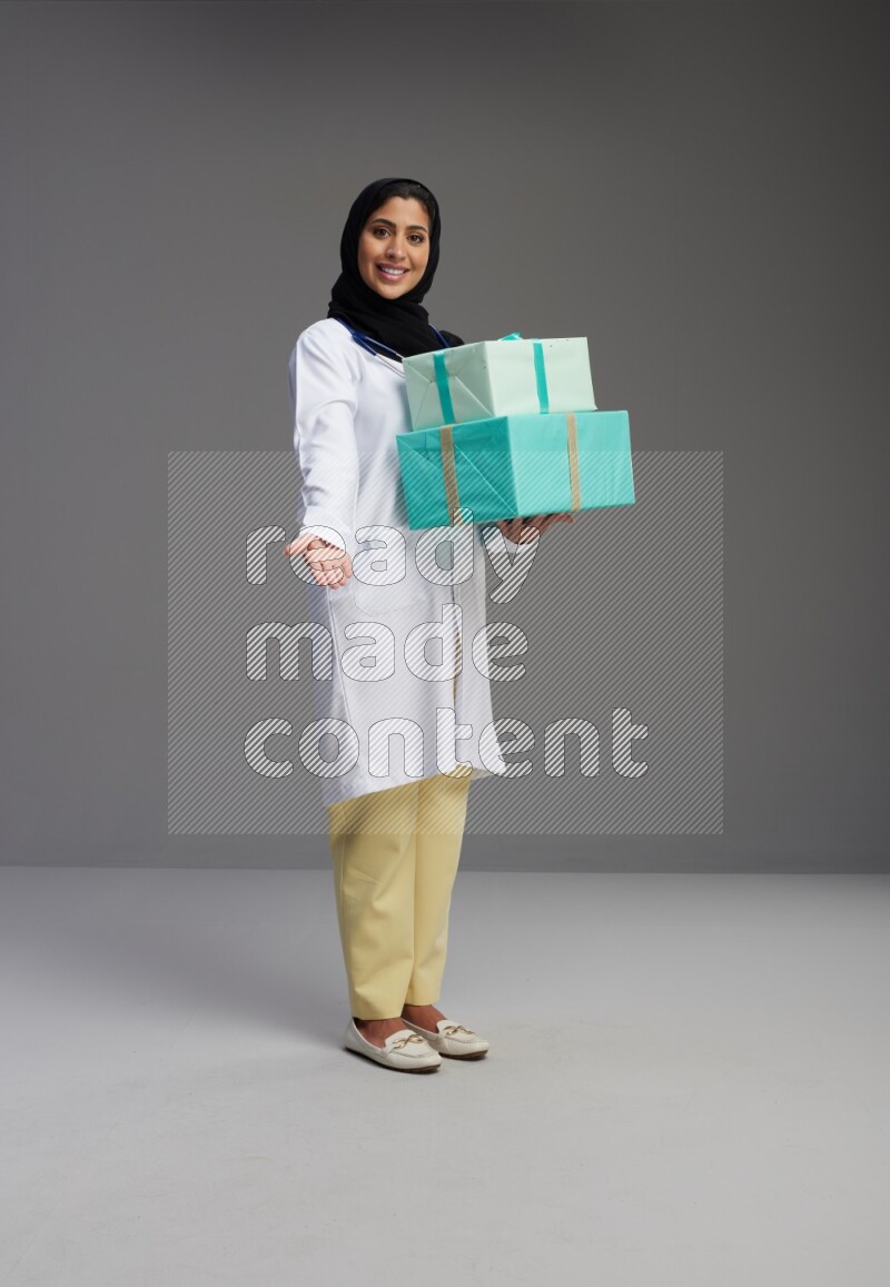 Saudi woman wearing lab coat with stethoscope standing holding gift box on Gray background