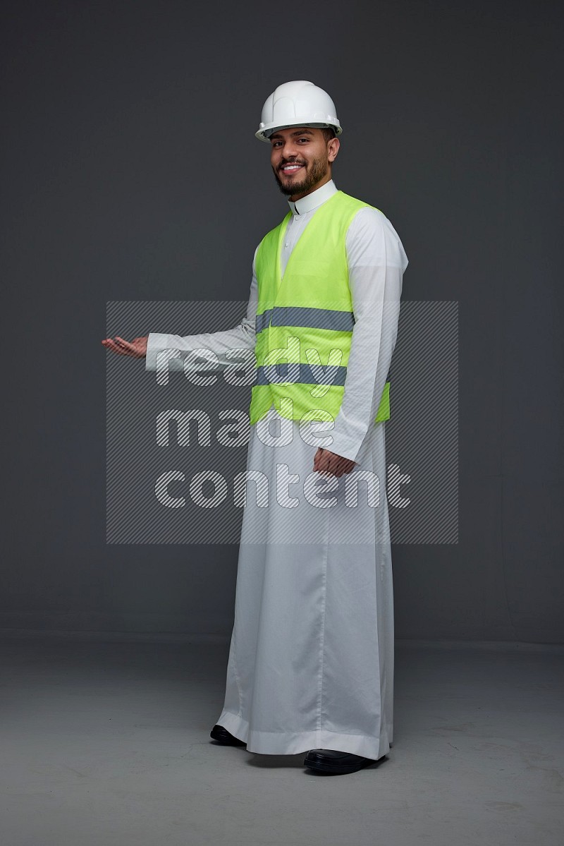 A Saudi man wearing Thobe with a yellow safety vest and white helmet standing and pointing different angles eye level on a gray background