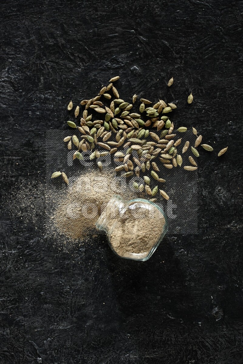 A flipped glass spice jar full of cardamom powder and powder spilled out of it with cardamom seeds spreaded on textured black flooring