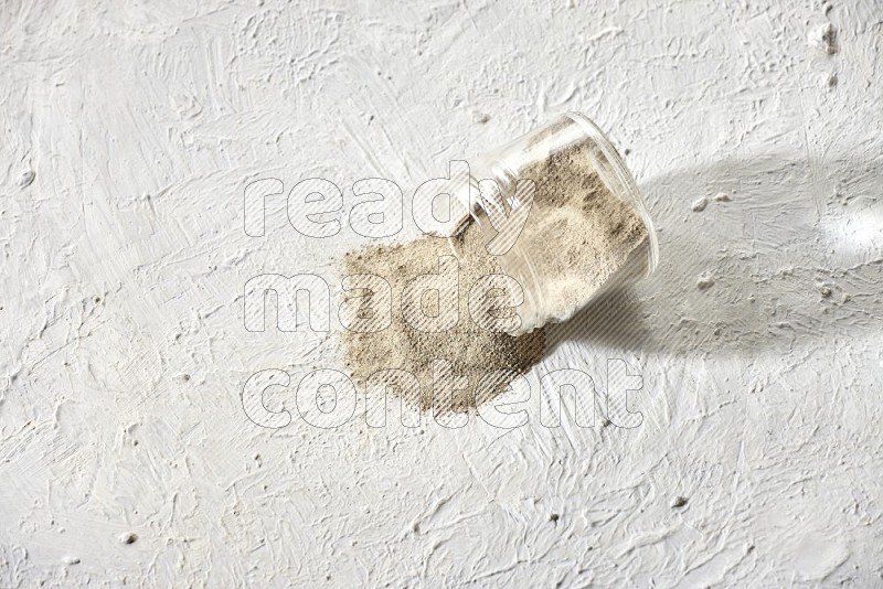 A flipped glass jar full of white pepper powder with spilled powder on textured white flooring