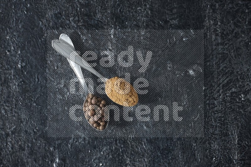 ملعقتان معدنيتان ممتلئتان ببودرة بهار الفلفل الحلو وحبوبه على خلفية سوداء