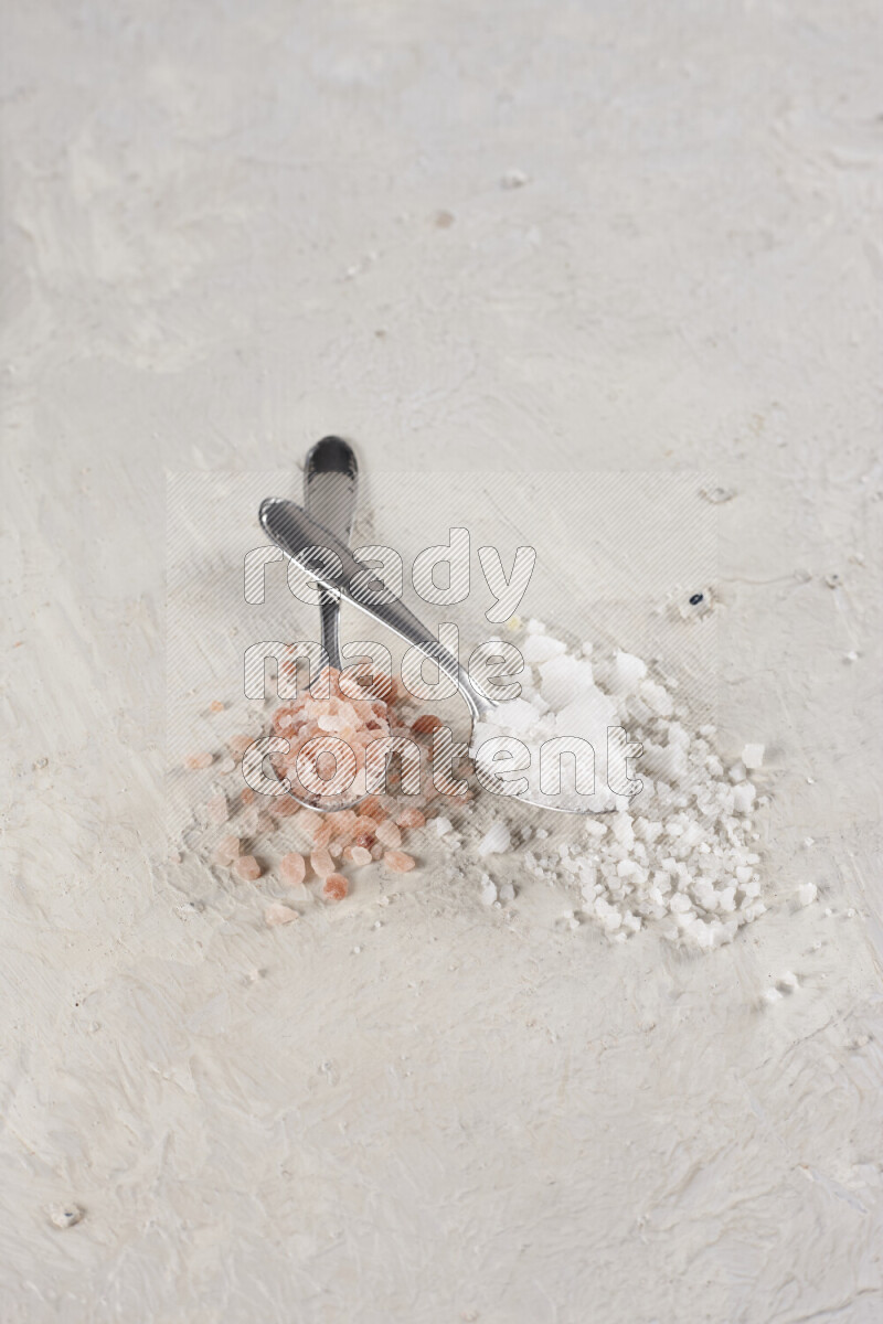2 metal spoons filled with white salt and pink himalayan salt on white background