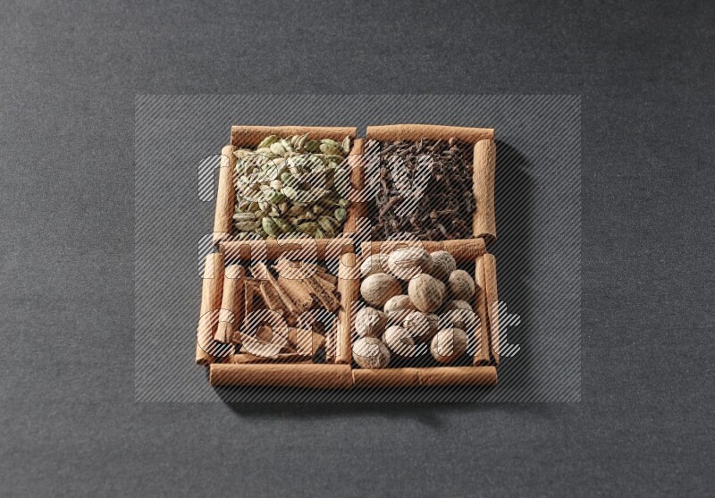 4 مربعات من عيدان القرفة ممتلئة بقطع القرفة وحبوب الهيل والقرنفل وجوزة الطيب على أرضية سوداء