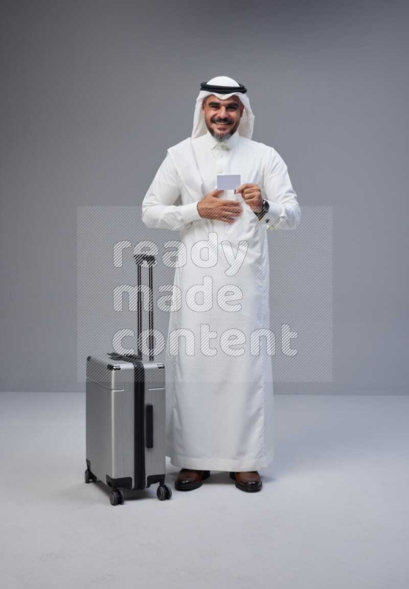 Saudi man wearing Thob and white Shomag standing holding Travel bag and ATM card on Gray background