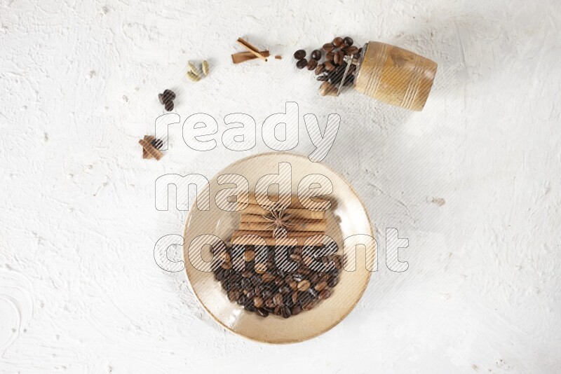 Beige plate full of coffee beans, cinnamon sticks and star anise with a coffee grinder, coffee beans, cinnamon pieces and cardamom next of it on white background