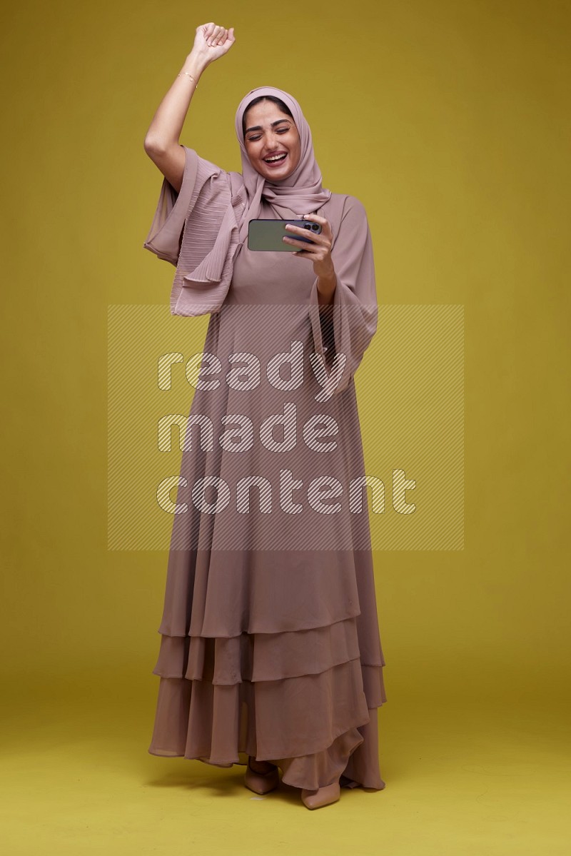 A woman Playing Games a Yellow Background wearing Brown Abaya with Hijab