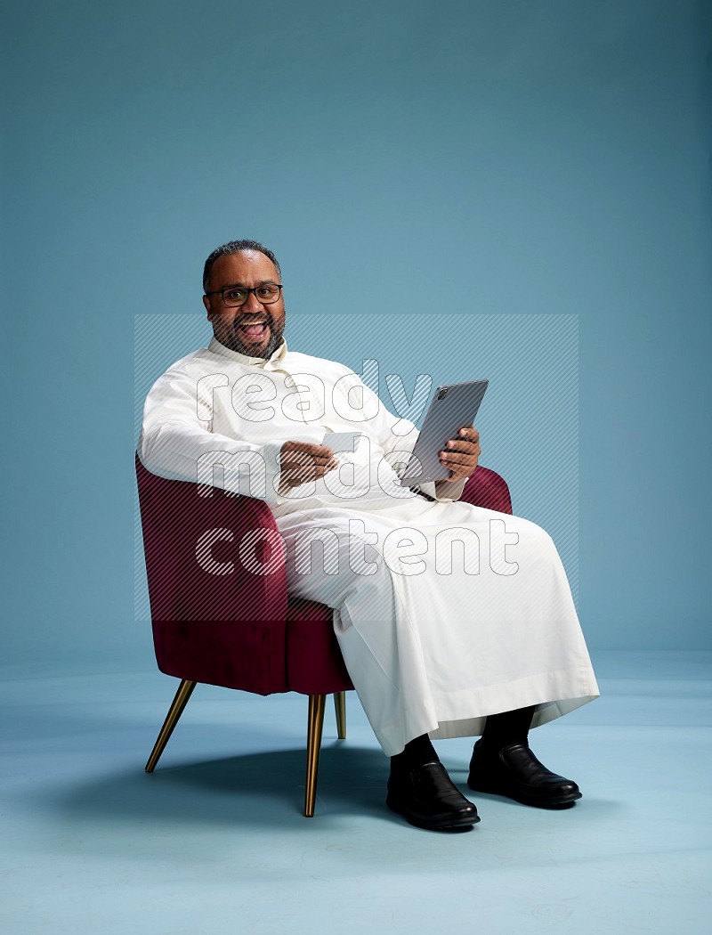 Saudi Man without shimag sitting on chair holding ATM card while working on tablet on blue background