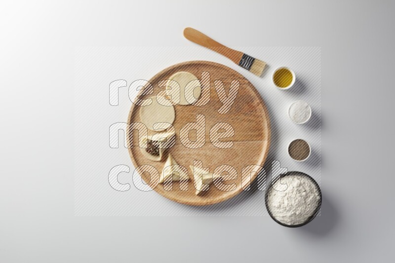 two closed sambosas and one open sambosa filled with meat while flour, salt, black pepper and oil with oil brush aside in a wooden dish on a white background