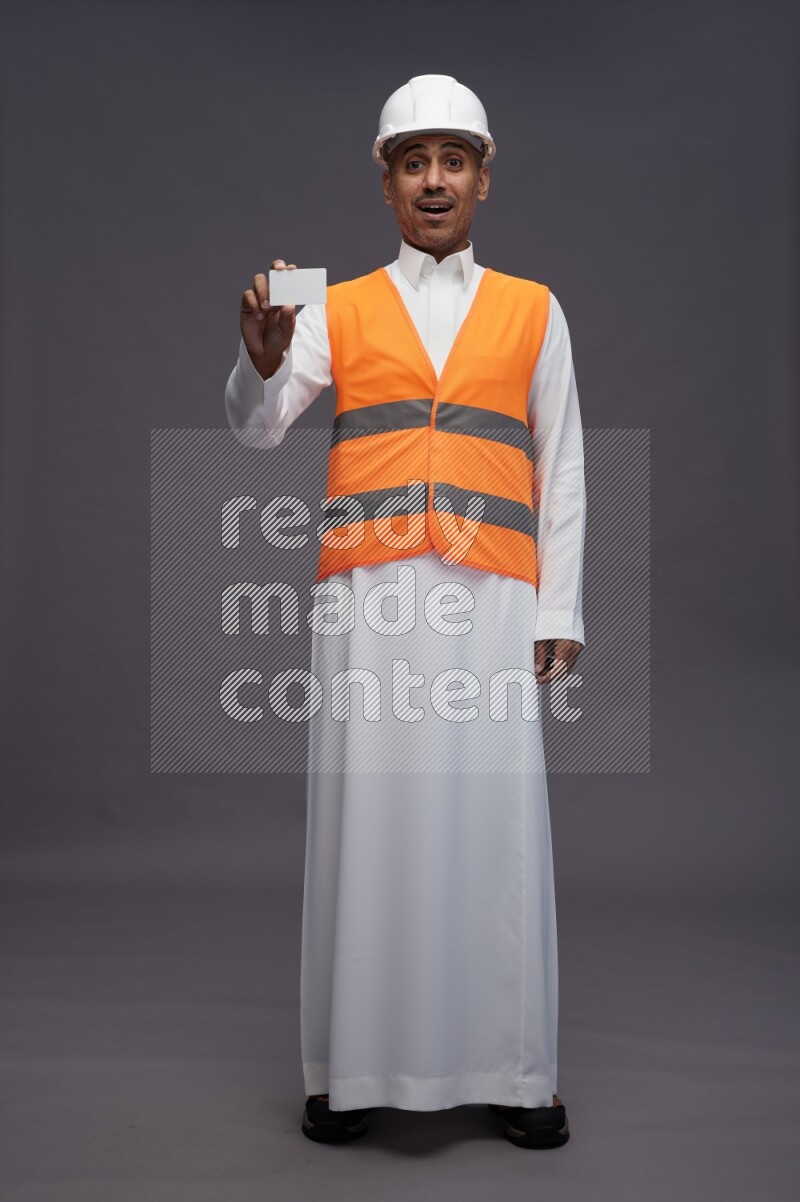 Saudi man wearing thob with engineer vest standing holding ATM card on gray background