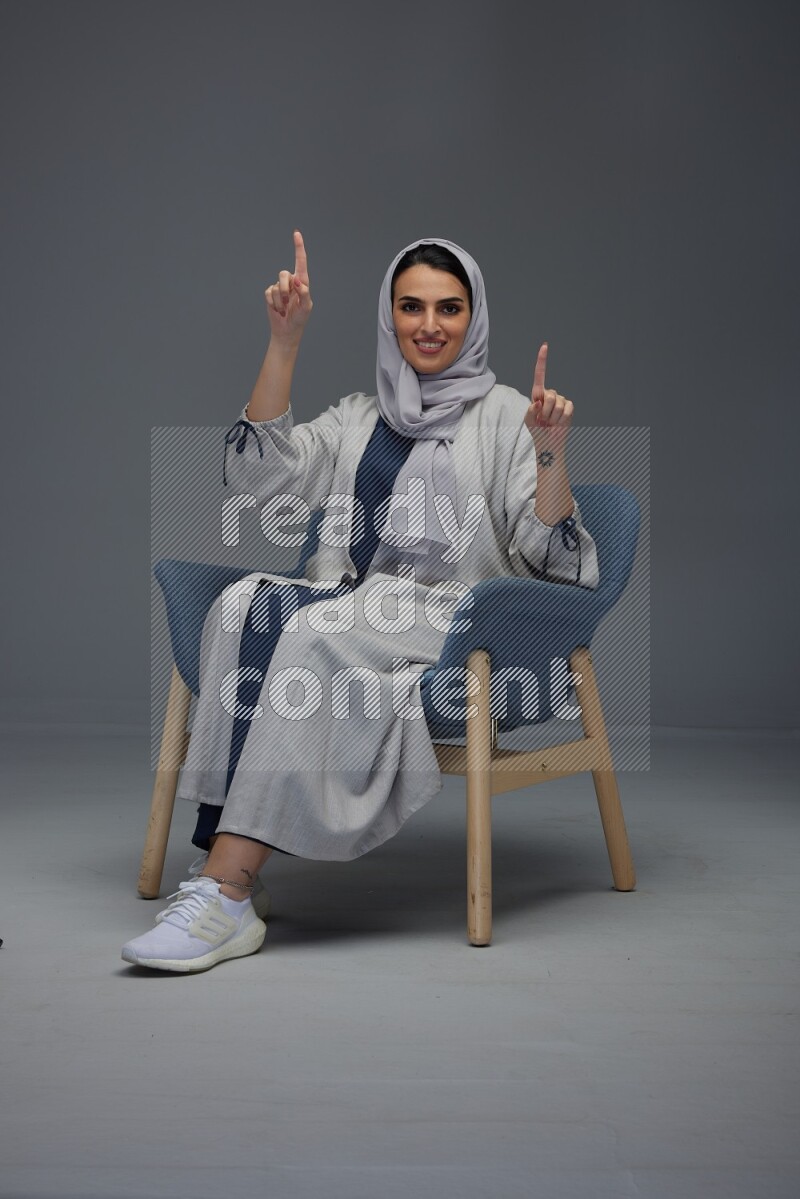 A Saudi woman wearing a light gray Abaya and head scarf sitting on a dark grey chair while making multi hand gestures eye level on a grey background