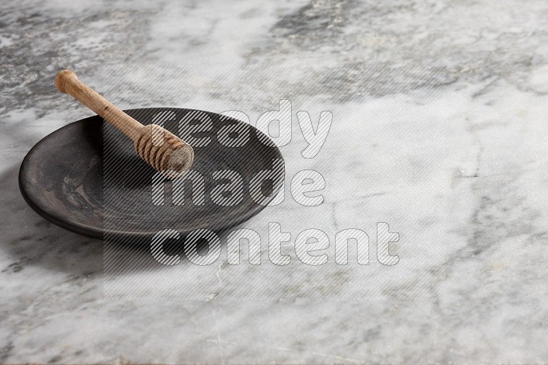 Black Pottery Plate with wooden honey handle in it, on grey marble flooring, 45 degree angle