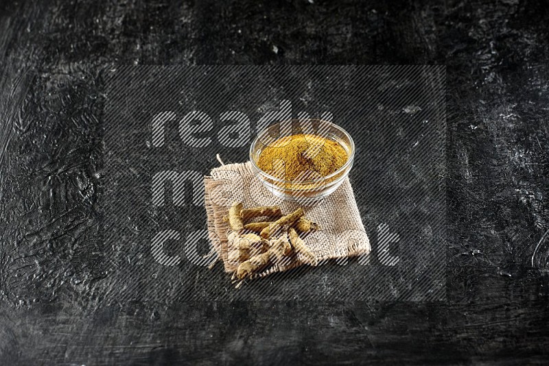 A glass bowl full of turmeric powder with dried turmeric fingers on a burlap fabric on textured black flooring