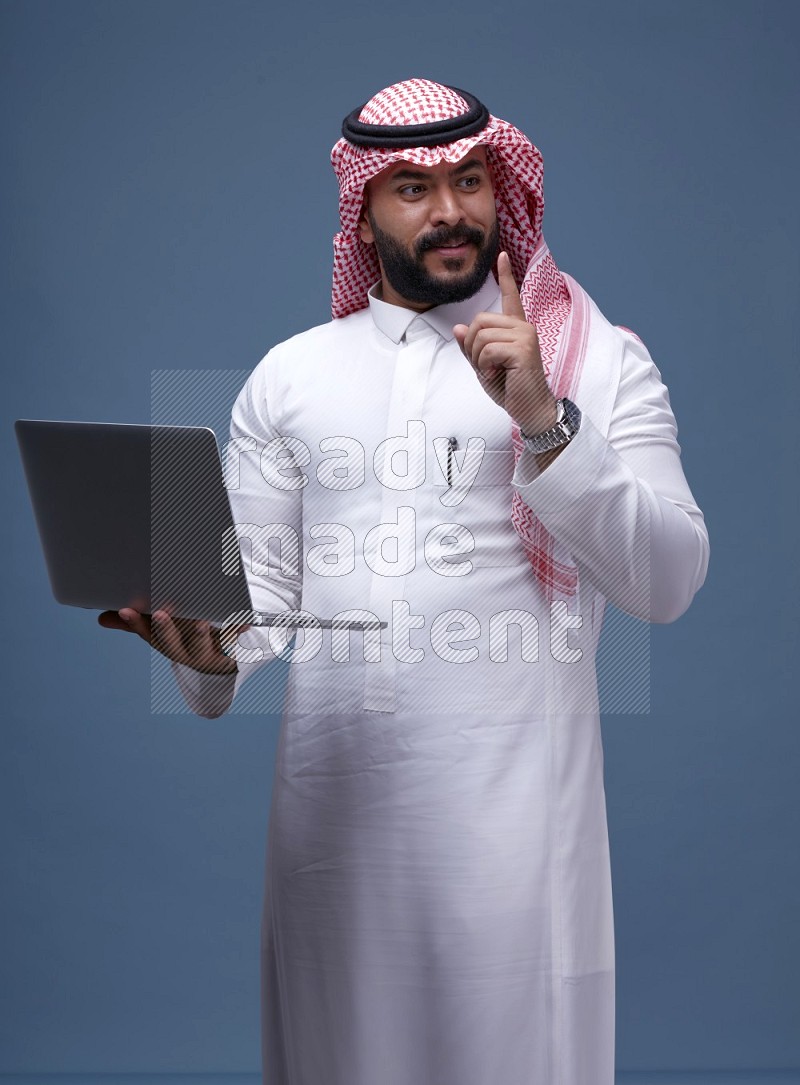 A man standing  with a laptop on Blue Background wearing Saudi Thob and Shomag