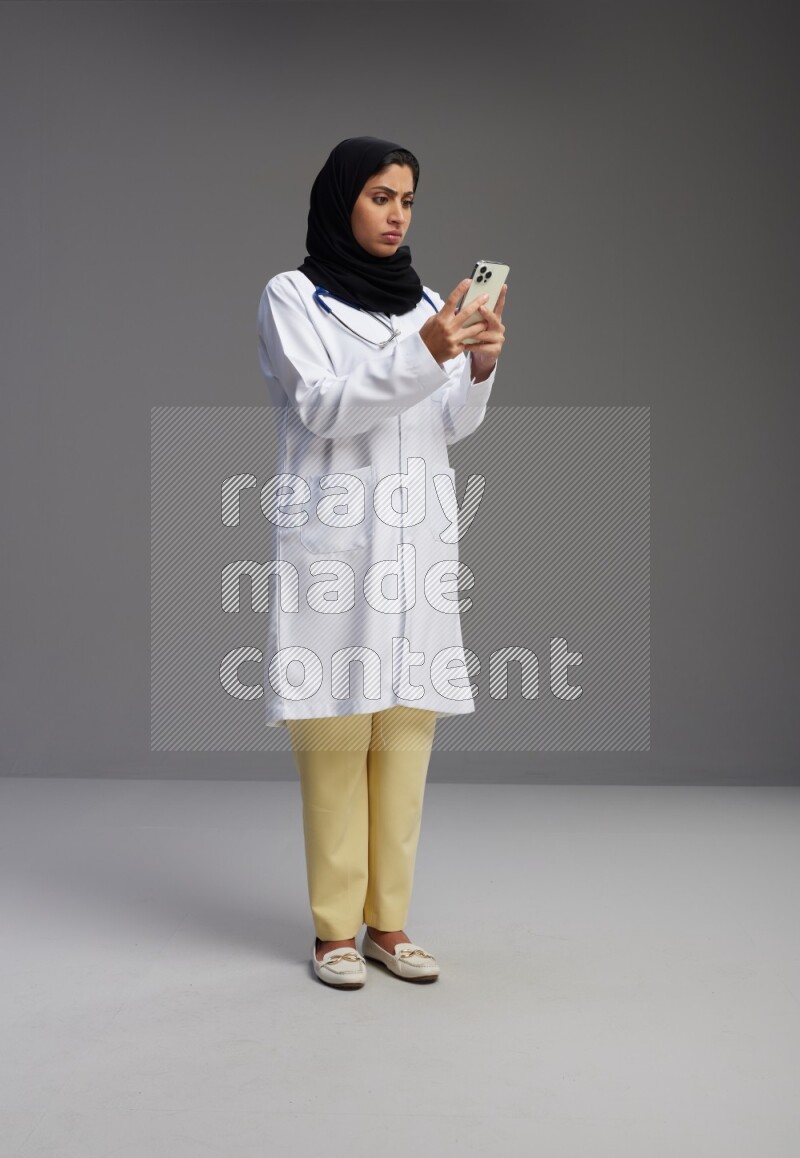 Saudi woman wearing lab coat with stethoscope standing texting on phone on Gray background