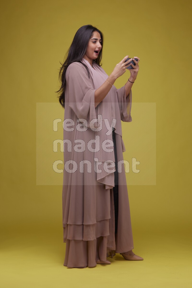 A woman Playing a Game on her smartphone on a Yellow Background wearing Brown Abaya
