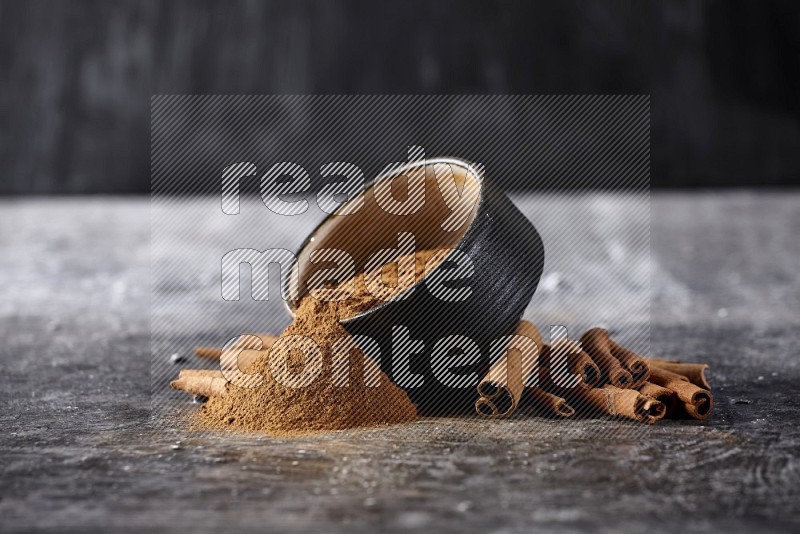 Black pottery bowl over filled with cinnamon powder and cinnamon sticks around the bowl on a textured black background