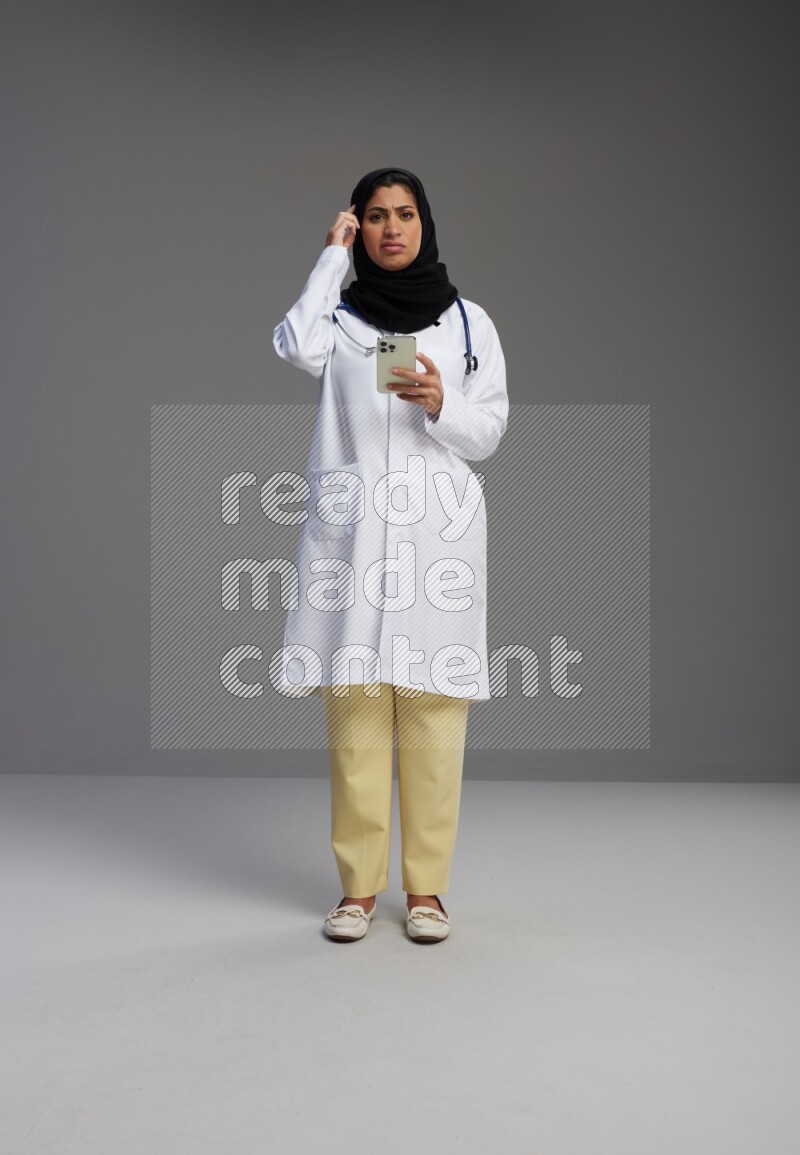 Saudi woman wearing lab coat with stethoscope standing texting on phone on Gray background