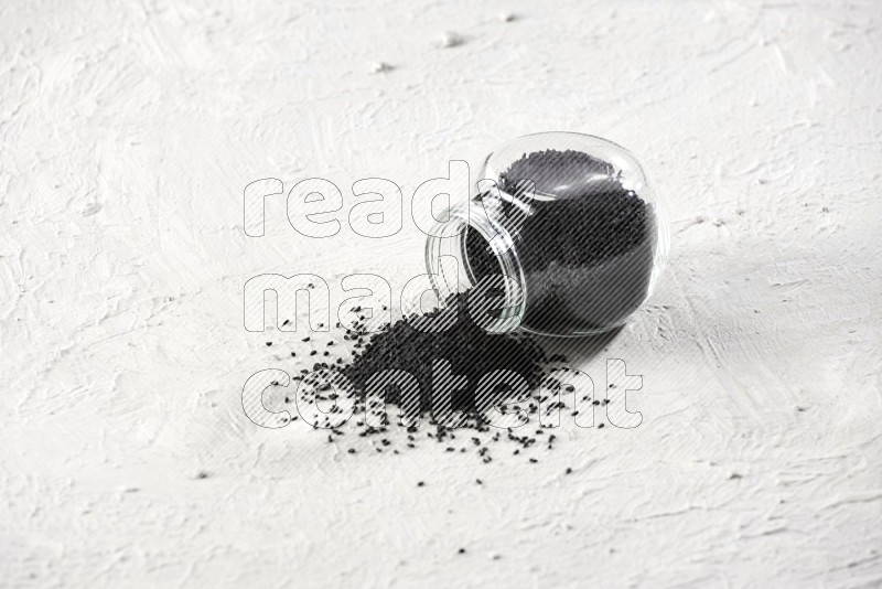 A glass spice jar full of black seeds and the jar flipped and seeds spread on a textured white flooring