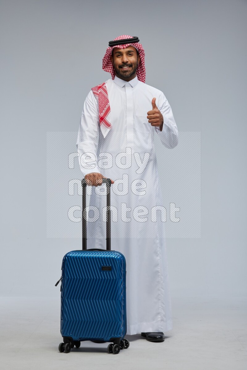 Saudi man wearing Thob and shomag standing holding Travel bag on Gray background