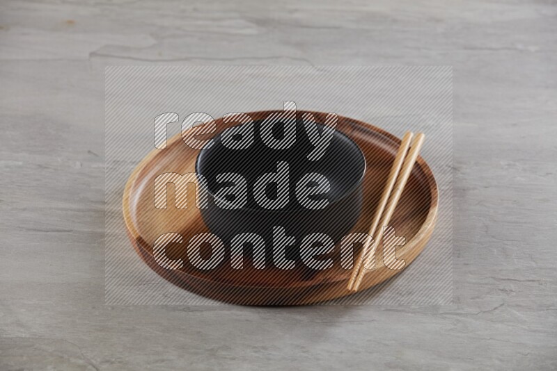 black ceramic round bowl on top of brown wood round plate and wood chopsticks, on grey textured countertop